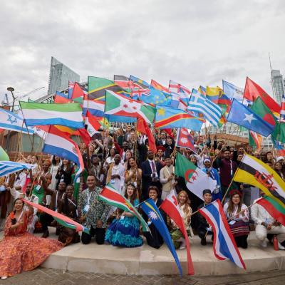 A large group of One Young World delegates are sitting and standing waving their home nation flags outside a conference venue,